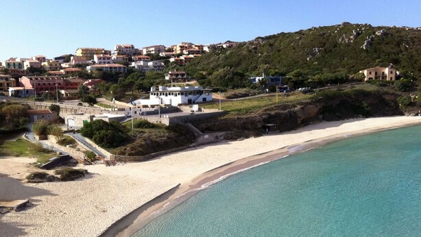 A coastal area with a sandy beach, clear turquoise waters, and a hillside with residential buildings. - Apartment in Santa Teresa Near the Beach (Santa Teresa di Gallura)