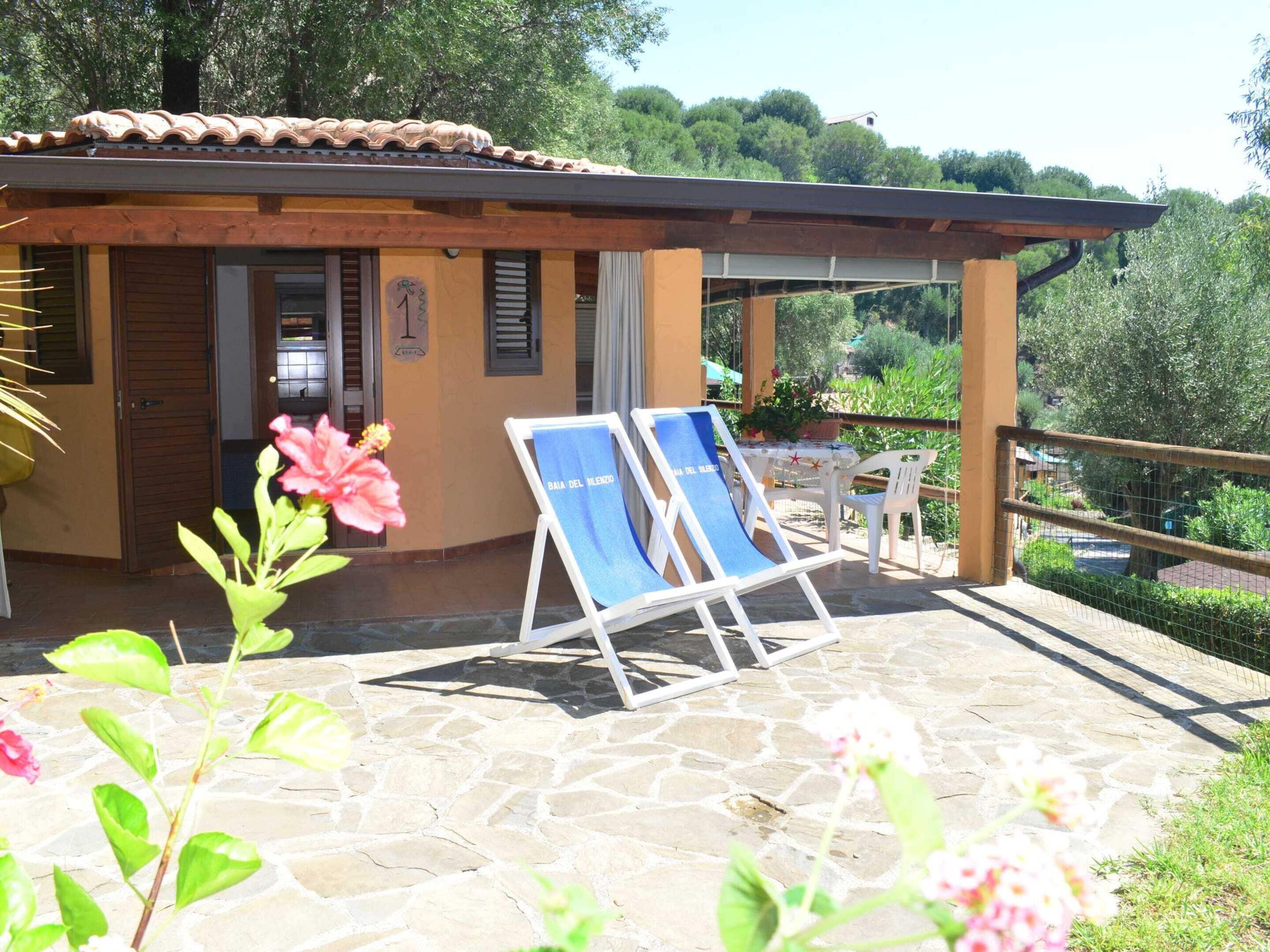 A small building with a tiled roof, a door, and a window. There are two lounge chairs with a towel on them, a table with a plant, and a chair. The building is surrounded by greenery and a stone floor.