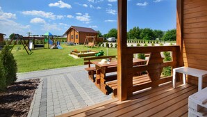 En træterrasse med et picnicbord og bænke, et legeområde med gyngestativ og rutchebane, samt udsigt til et hus og træer i baggrunden.