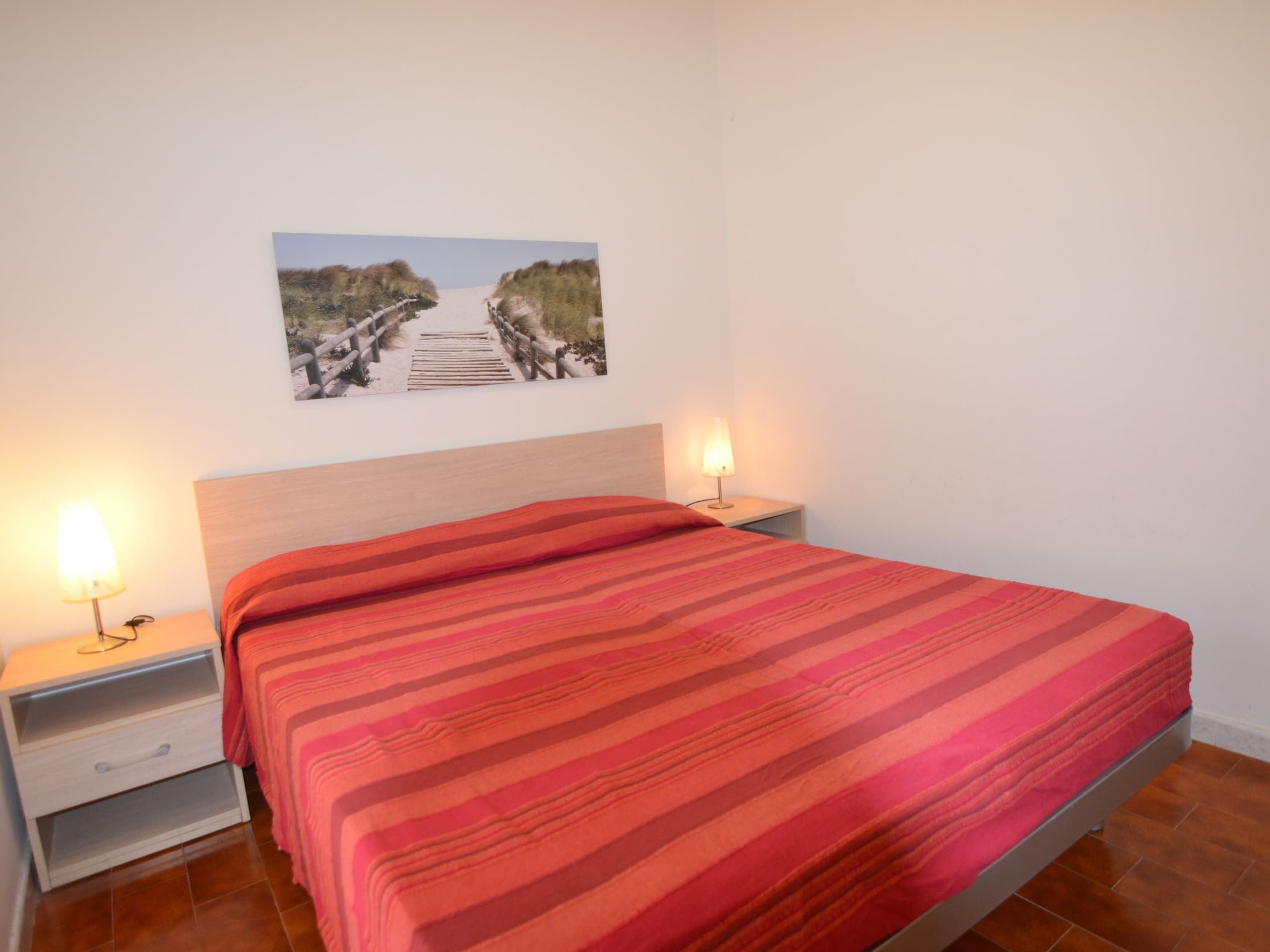 A bedroom with a bed, two bedside tables, a nightstand, a lamp, a picture on the wall, and a wooden headboard.
