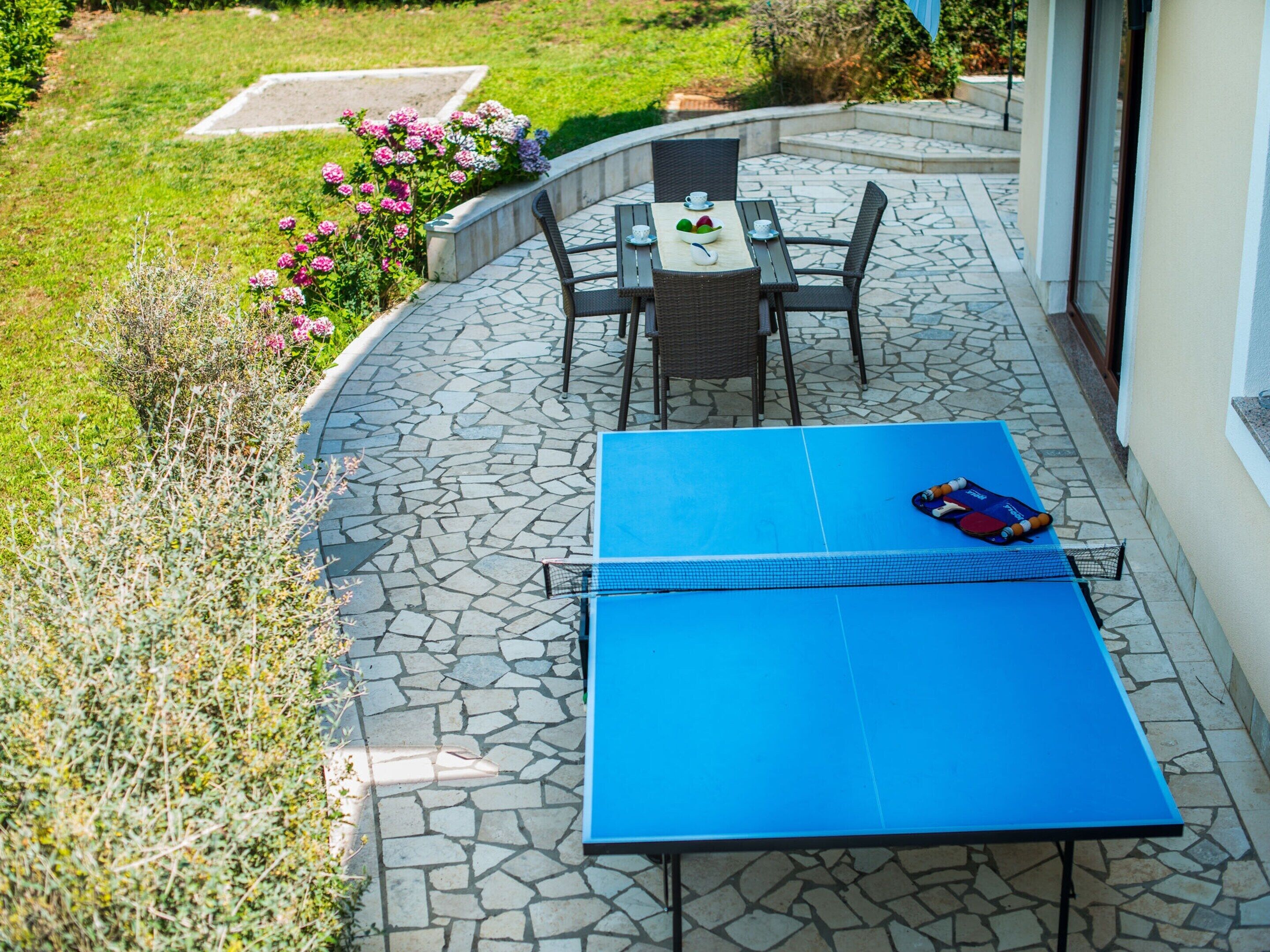 Eine Terrasse mit blauem Tisch und Stühlen, einem Badmintonnetz und einem Blumengarten.
