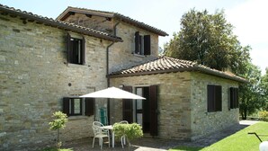 Un edificio in pietra con tetto a tegole, un patio con tavolo e sedie, e un piccolo albero.