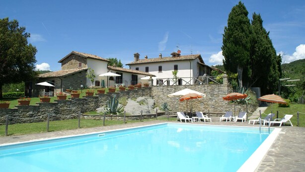 Una piscina con lettini e ombrelloni, circondata da muri in pietra e un edificio con tetto in tegole.
