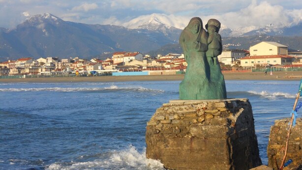 Un pueblo costero con una estatua de dos figuras sobre una roca, una cadena montañosa al fondo y edificios a lo largo de la orilla.