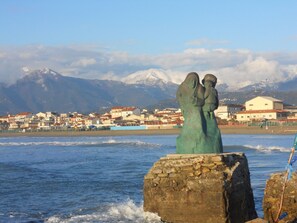 Una località costiera con una statua di due figure su una roccia, una catena montuosa sullo sfondo e edifici lungo la riva.