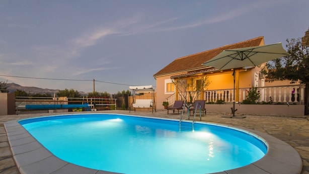 A swimming pool with a stone border, a patio area with a swing set, and a house with a tiled roof in the background. - Holiday Home Near Zadar With Private Pool (Posedarje)