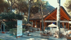 An outdoor restaurant with wooden tables and umbrellas, surrounded by trees and a mountain in the background. - Bungalow in Po Delta Park With Beach Access (Comacchio)