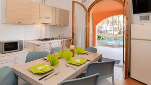 A modern kitchen with wooden cabinets, a gas stove, and a microwave. There is a dining table set with green placemats and silverware. An open door leads to an outdoor area with a view of a pool and lounge chairs. - Bungalow in Po Delta Park With Beach Access (Comacchio)