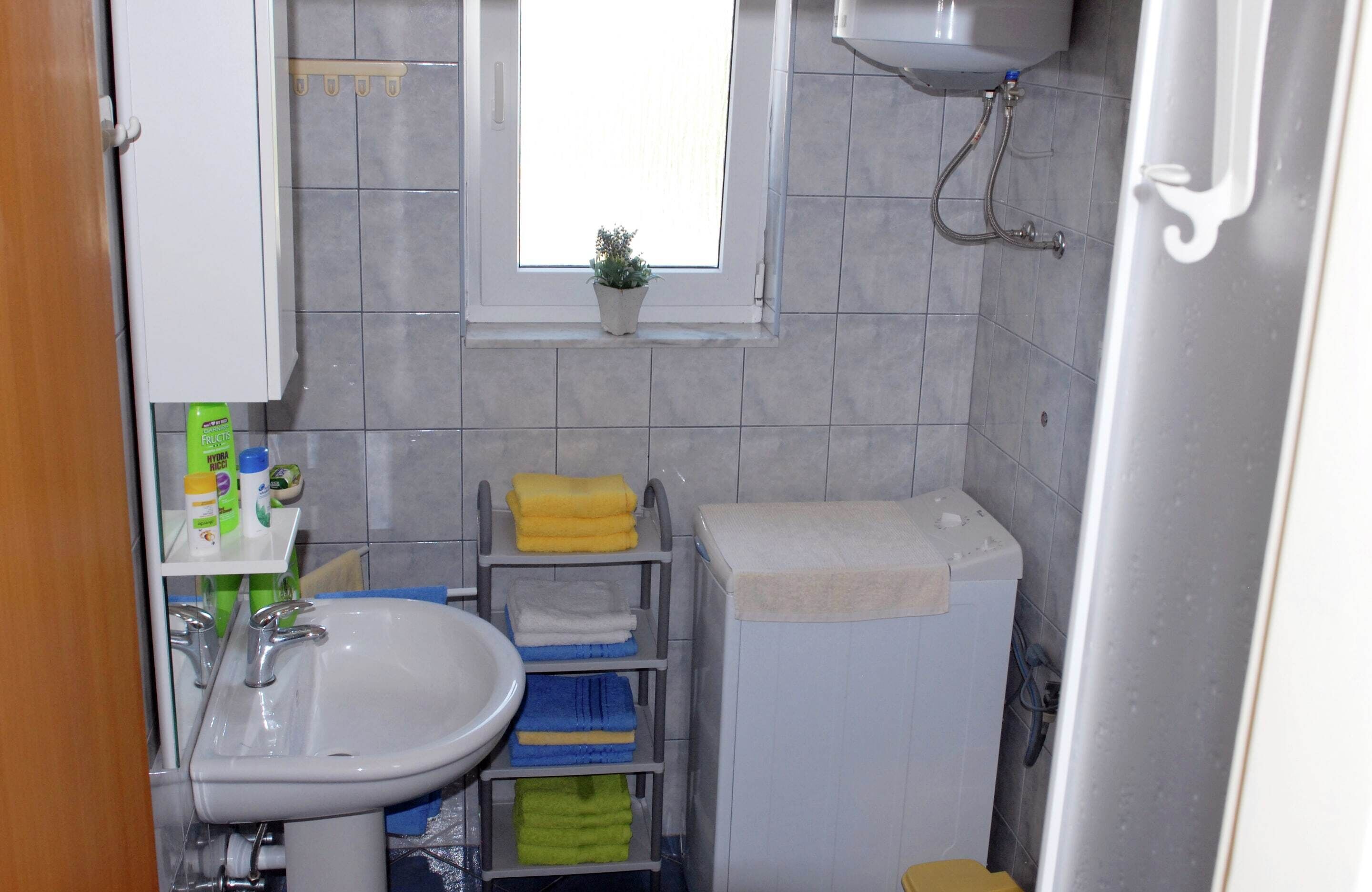 Une petite salle de bain avec un lavabo, des toilettes, une fenêtre avec une plante et une étagère contenant des articles de toilette.