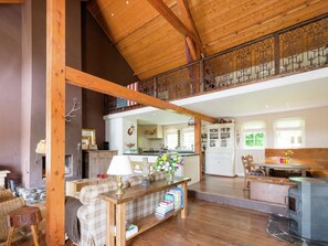 A spacious living area with a wooden ceiling, a fireplace, a sofa, a dining table with books, and a kitchen in the background. - Peace, Nature & Comfort Combined (Houffalize)