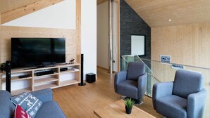 Un salon moderne doté d’un grand téléviseur à écran plat, de fauteuils recouverts d’un tissu gris, d’un plancher en bois et d’un escalier menant à un niveau supérieur.