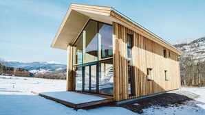 Une cabane moderne en bois, dotée de grandes fenêtres et d’un toit en pente, située dans un paysage enneigé avec des montagnes en arrière-plan.