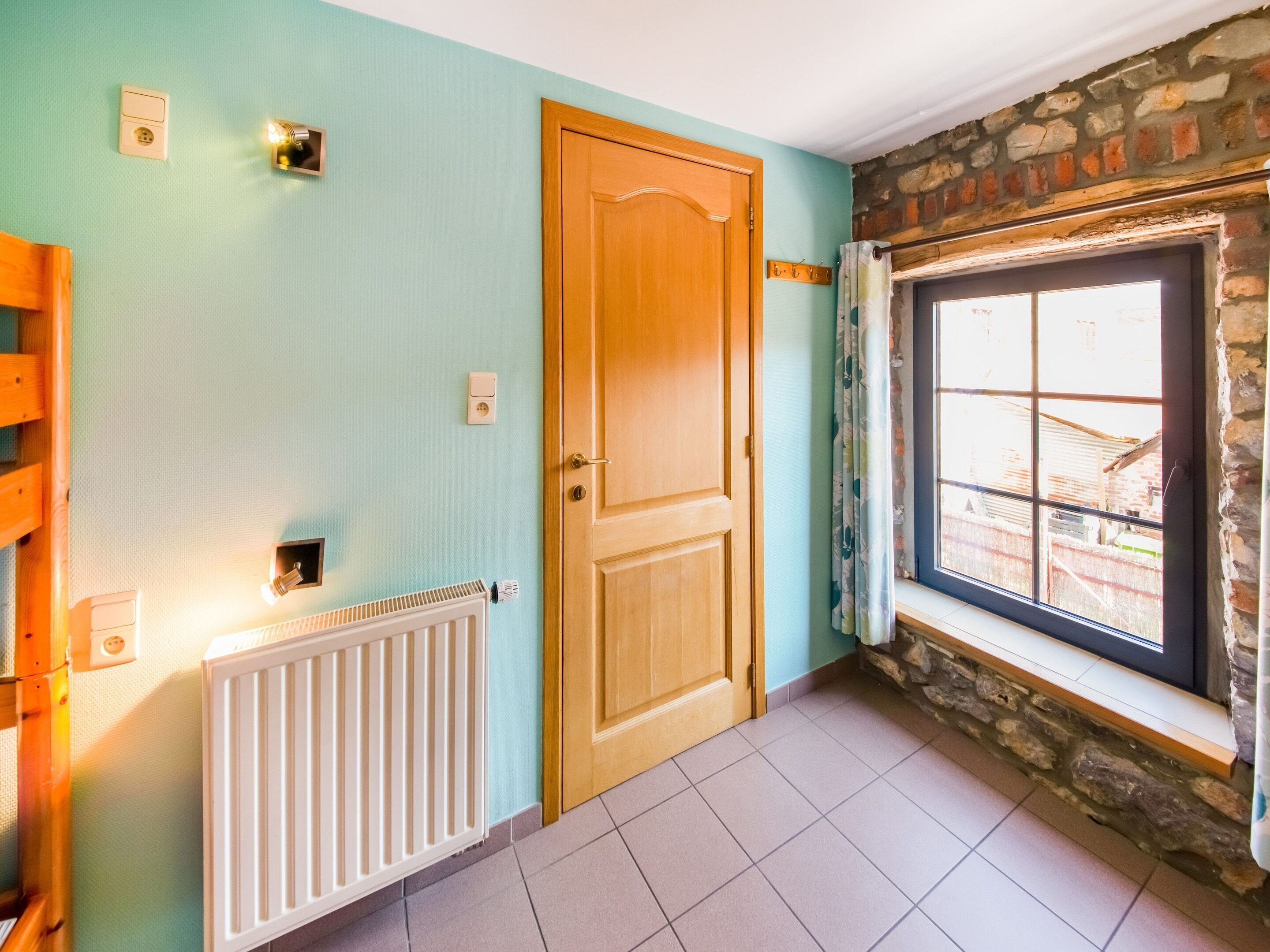 Una habitación con puerta de madera, ventana con vistas, radiador y un interruptor de luz en la pared.