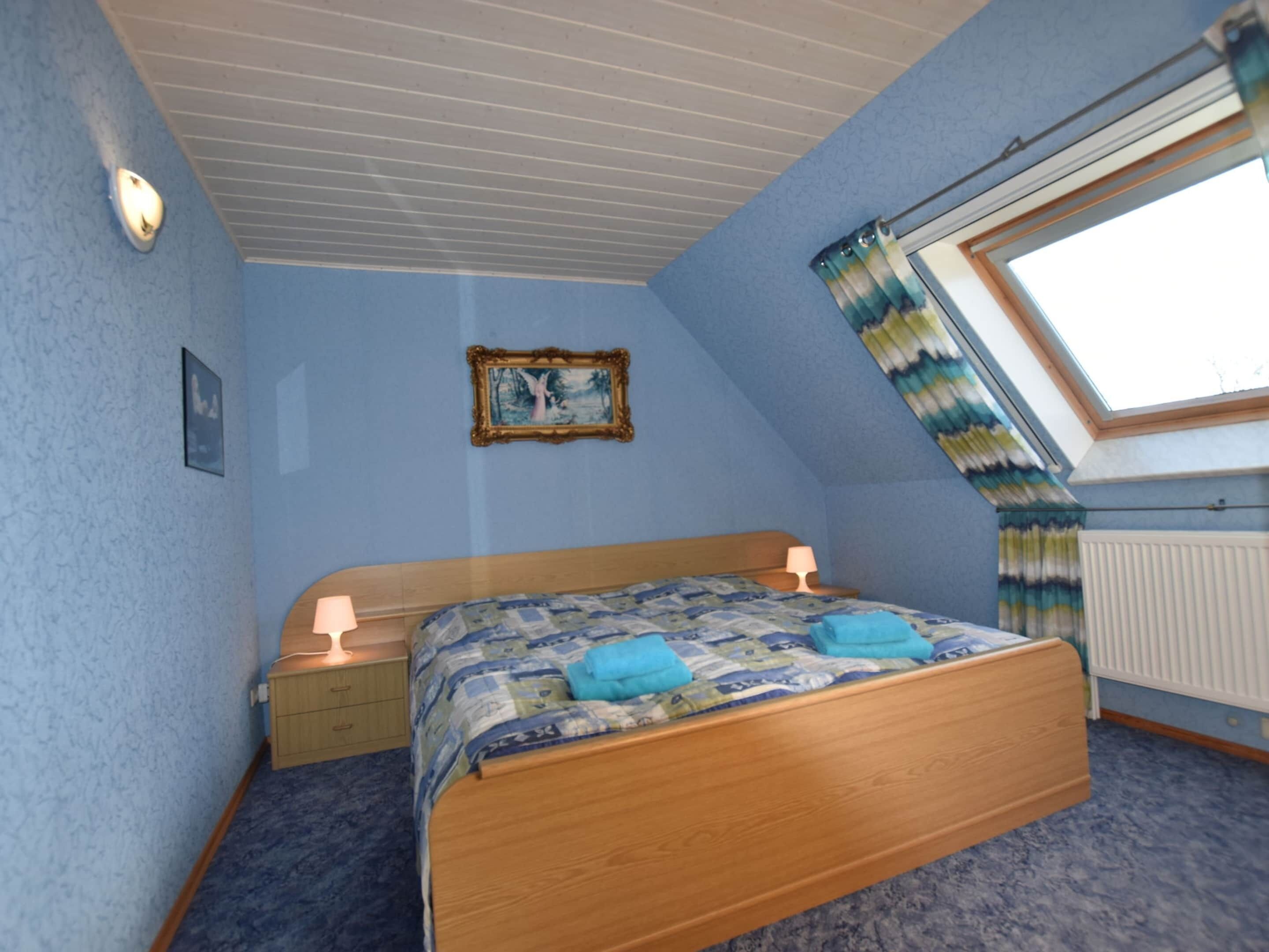 A bedroom with a wooden bed, a nightstand, a lamp, a framed picture on the wall, and a window with curtains.