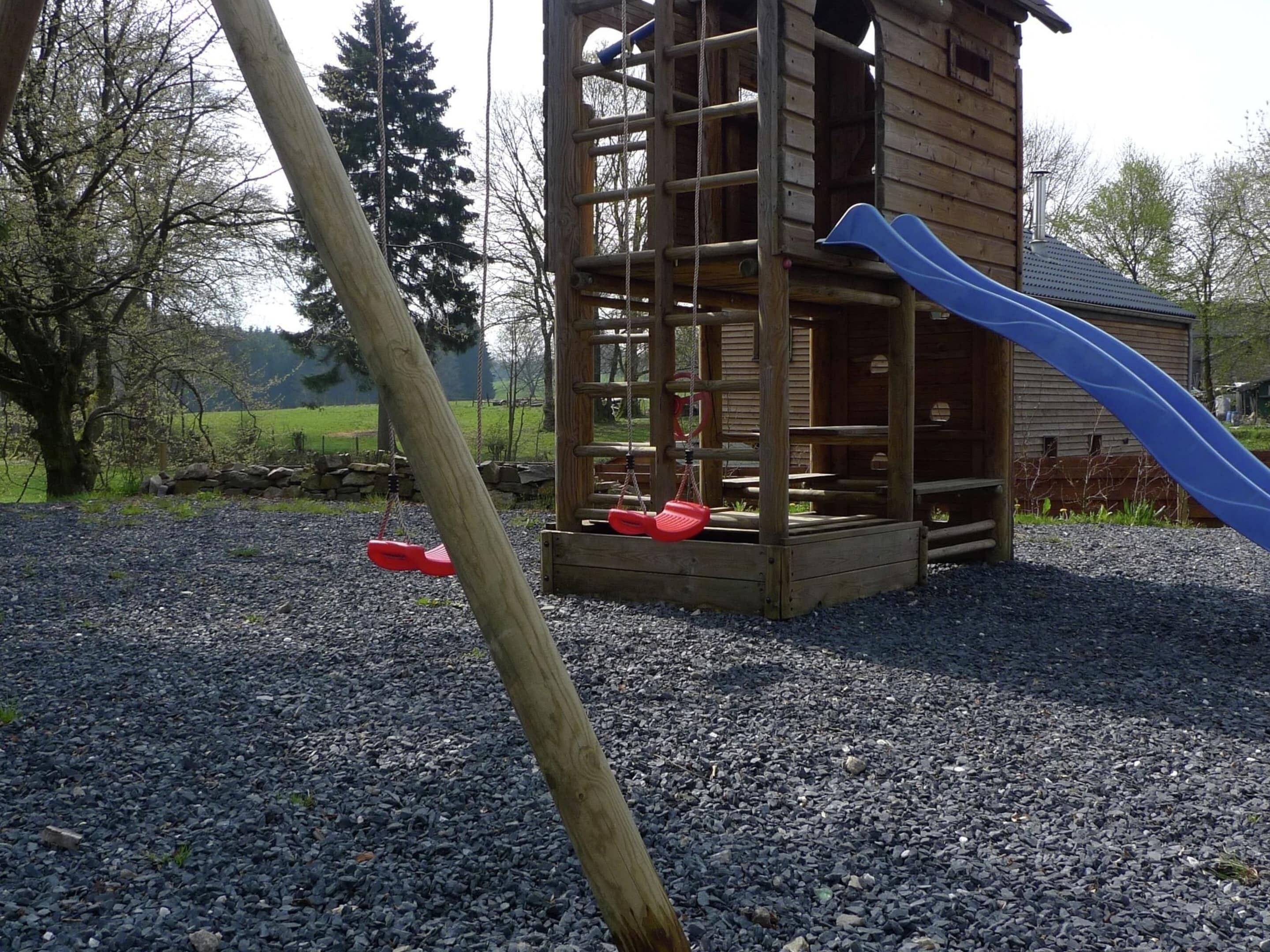 A wooden play structure with a slide and swings, set on a gravel surface with trees and a building in the background.
