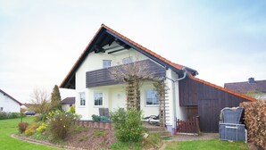 A two-story house with a sloped roof, a garden with various plants, and a wooden fence. - Ferienhaus in Hessen mit Grossem Garten (Voehl)