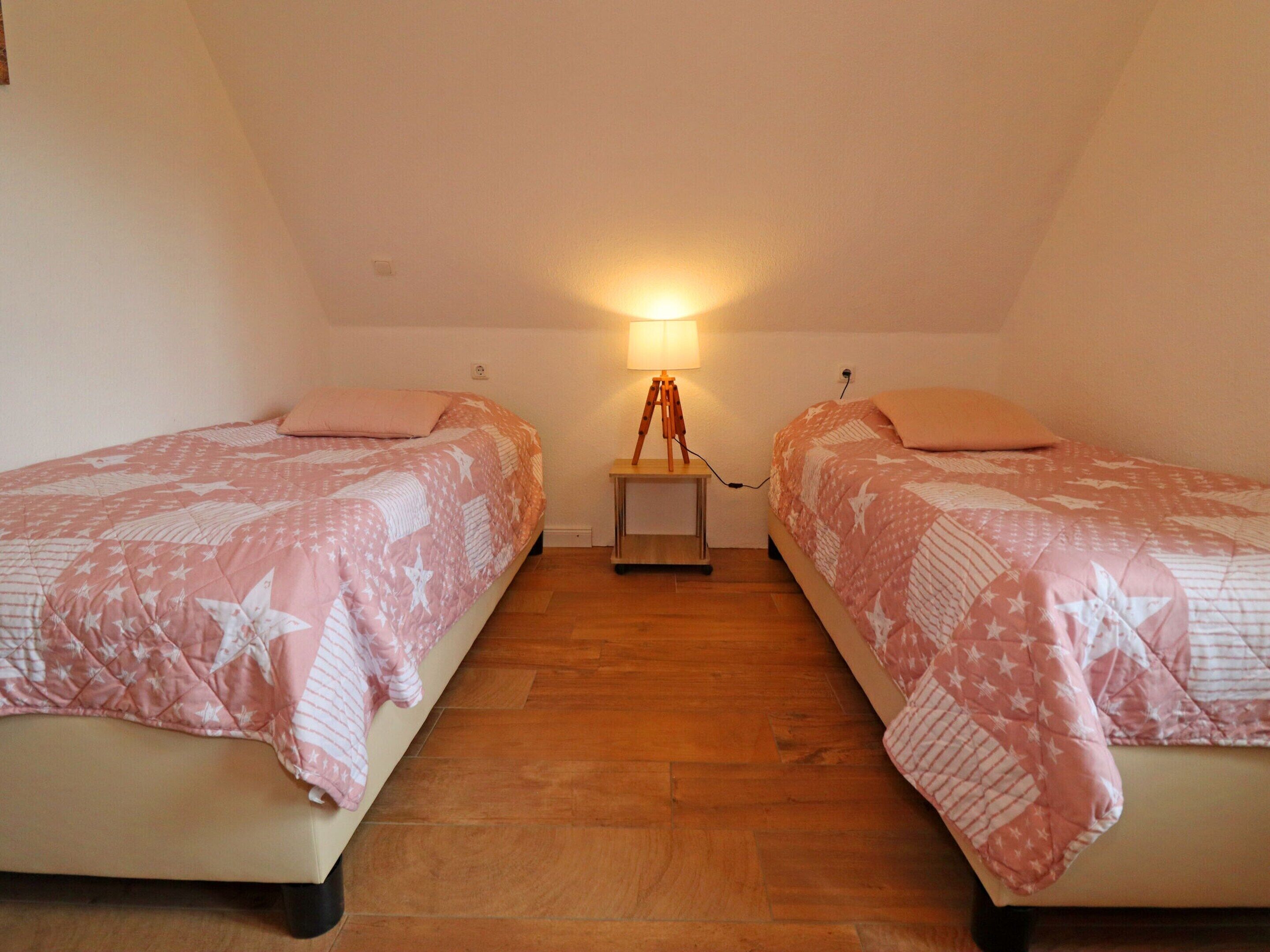 Ein Zimmer mit zwei Einzelbetten, jeweils mit einer rosa-weißen Bettdecke, einem Nachttisch mit Lampe und einem Holzboden.