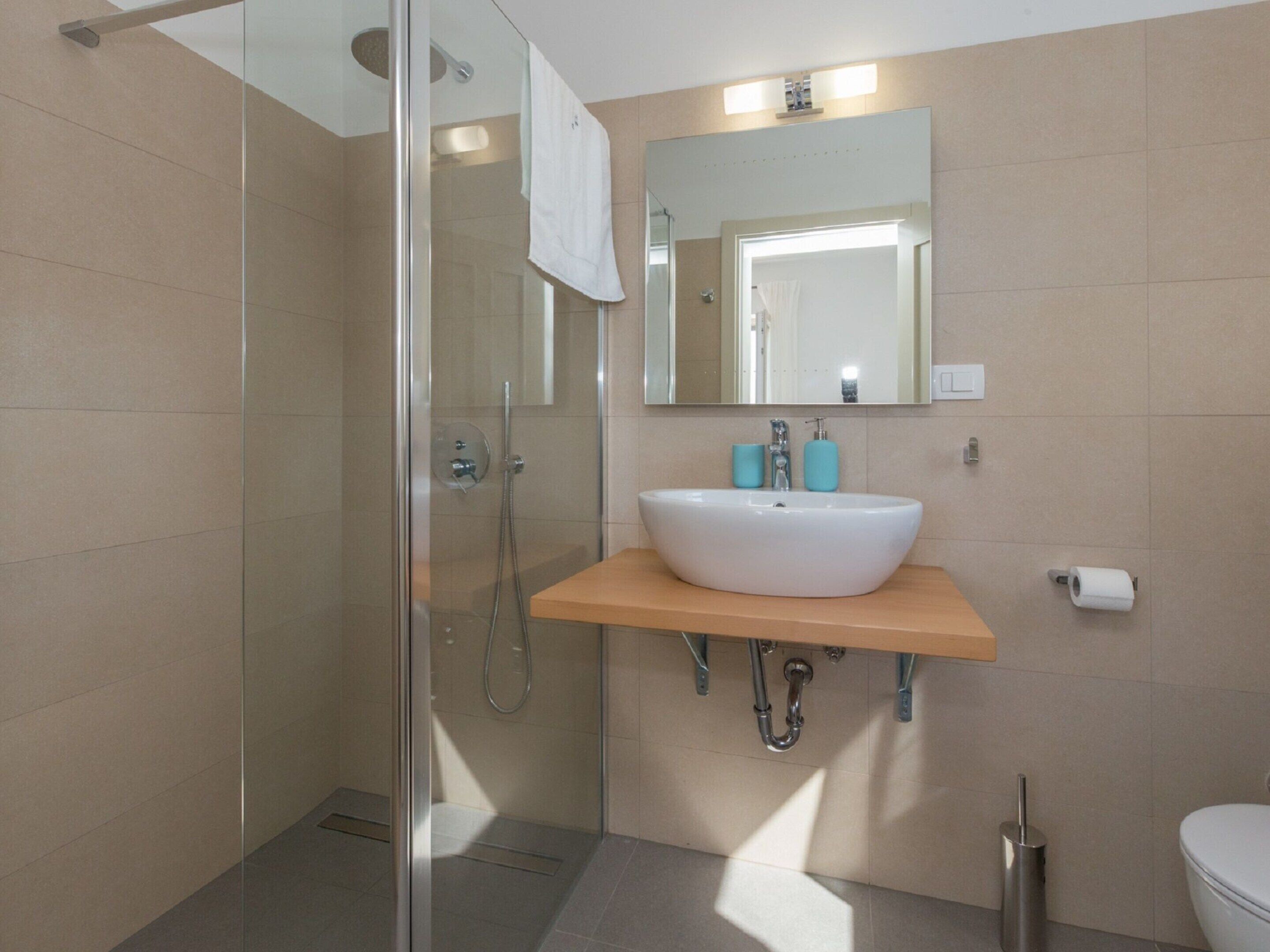 A modern bathroom with a glass shower enclosure, a white sink with a wooden countertop, a large mirror, and a toilet.