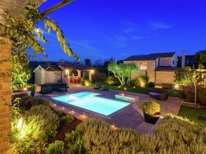 Un área de piscina exterior bien iluminada, con terraza, asientos y un pequeño jardín.