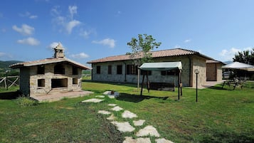 Um edifício de pedra com telhado em cerâmica, uma pequena construção com chaminé e uma mesa de piquenique com toldo.
