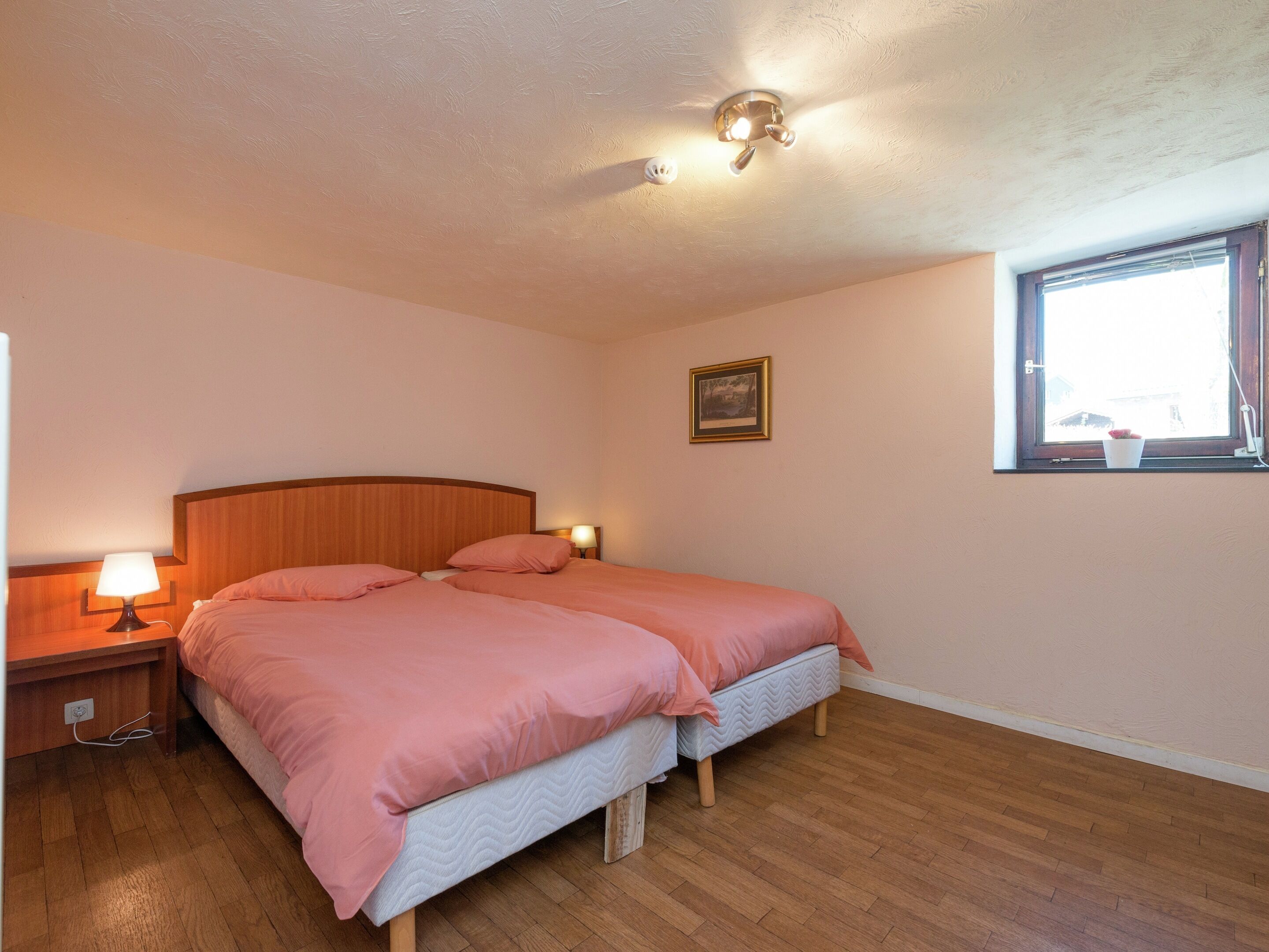Une chambre avec une tête de lit en bois, deux lits, une table de chevet avec une lampe, une fenêtre donnant sur un beau paysage et un luminaire au plafond.