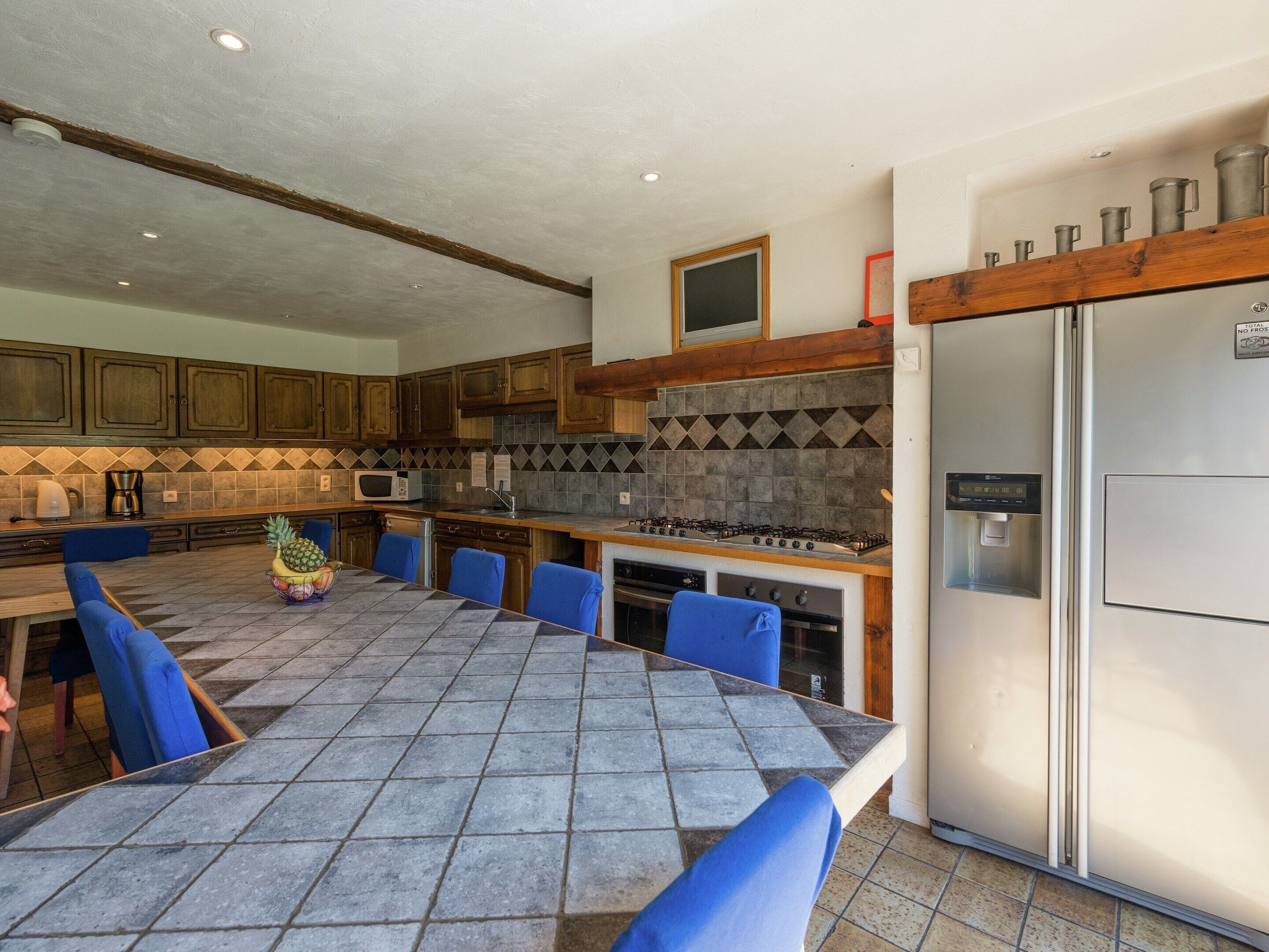 Uma cozinha com ilha revestida de azulejos, cadeiras azuis, geladeira de aço inoxidável e armários de madeira.