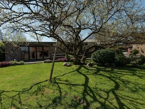 Een bakstenen gebouw met een grote boom op de voorgrond, een verzorgd gazon en een stenen muur op de achtergrond.