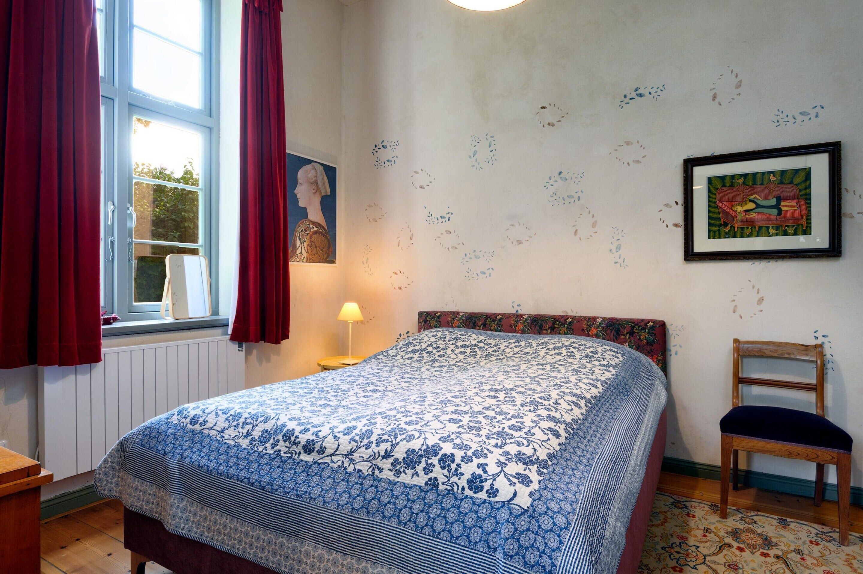 Ein Schlafzimmer mit Bett, Sessel, Fenster mit Vorhängen, Lampe und Wanddekorationen.