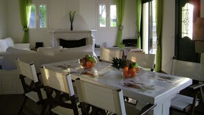 A dining area with a table set for four, surrounded by chairs. A fireplace is visible in the background, and there are windows with green curtains. - Comfy Apartment in Vinkuran With the Beach Nearby (Corfu)