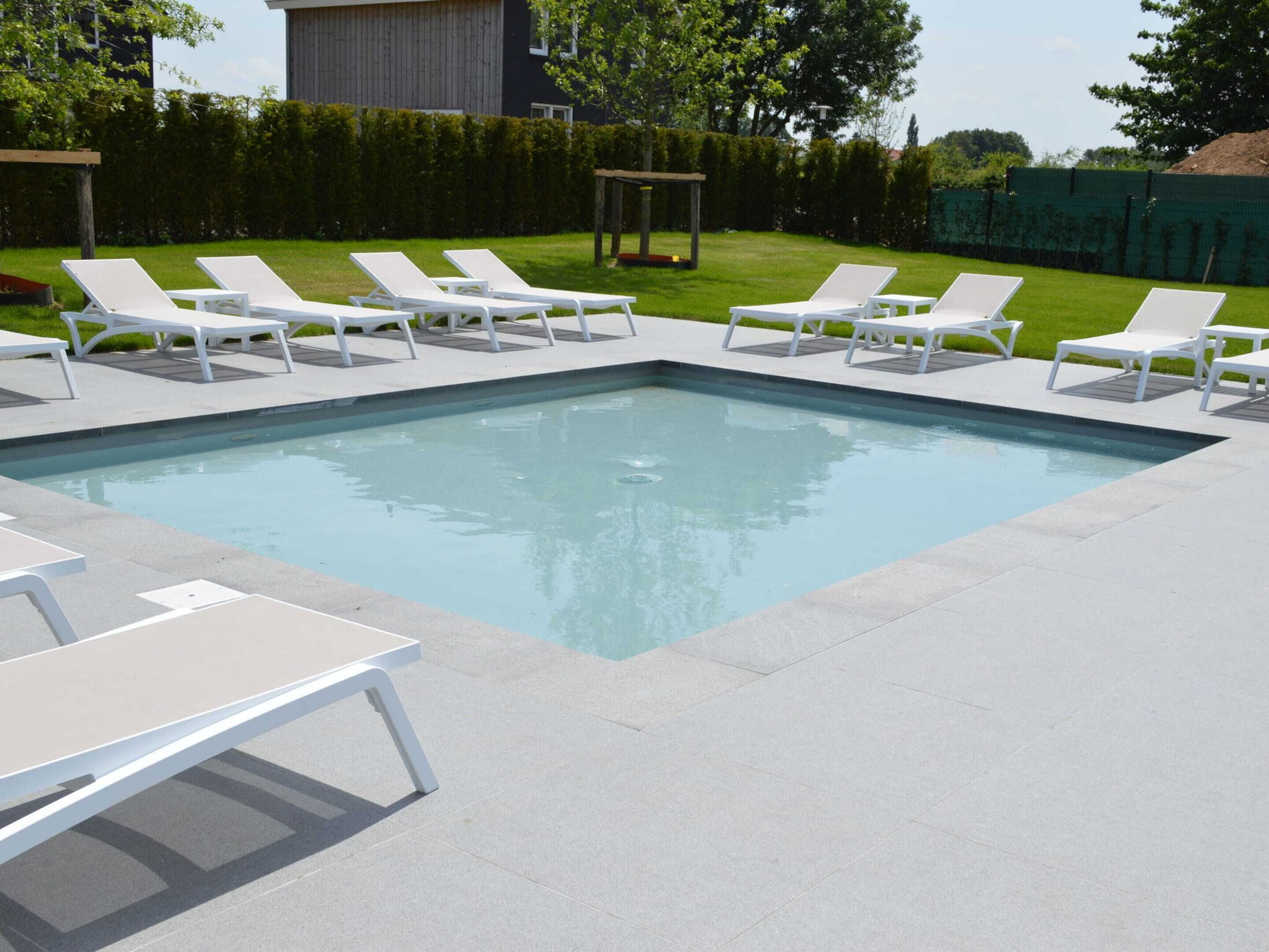 A rectangular swimming pool with white lounge chairs and tables arranged around it.