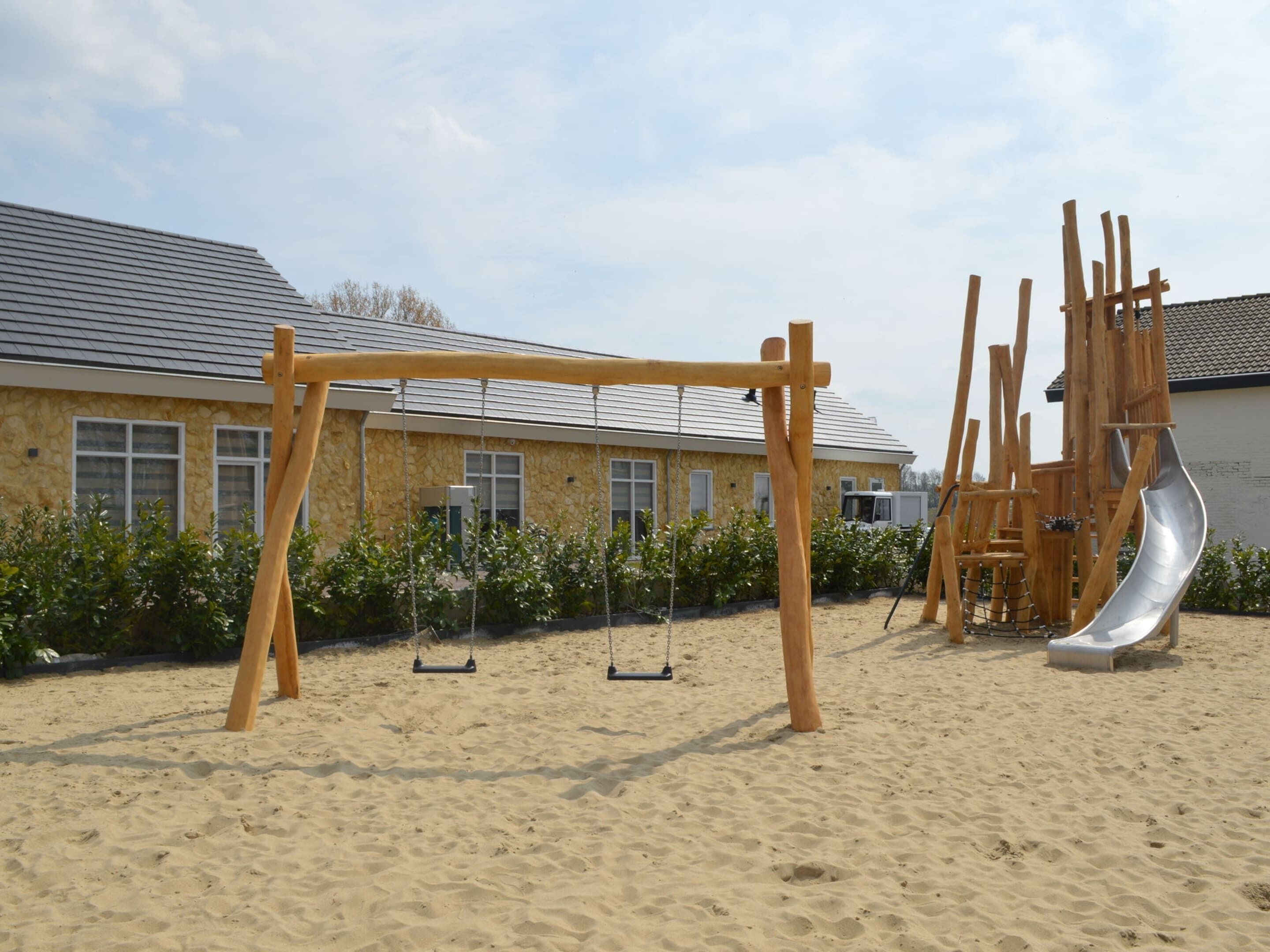 Un espacio al aire libre con arena, un conjunto de columpios de madera, un tobogán y un edificio de fondo con paredes de piedra y ventanas.
