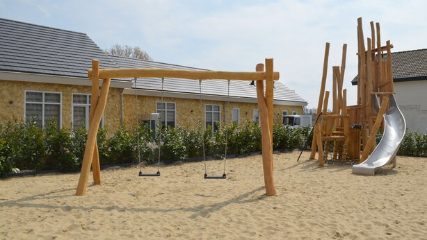 Ein sandiger Außenbereich mit einer Holz-Schaukel, einer Rutsche und einem Gebäude im Hintergrund mit Steinmauern und Fenstern.