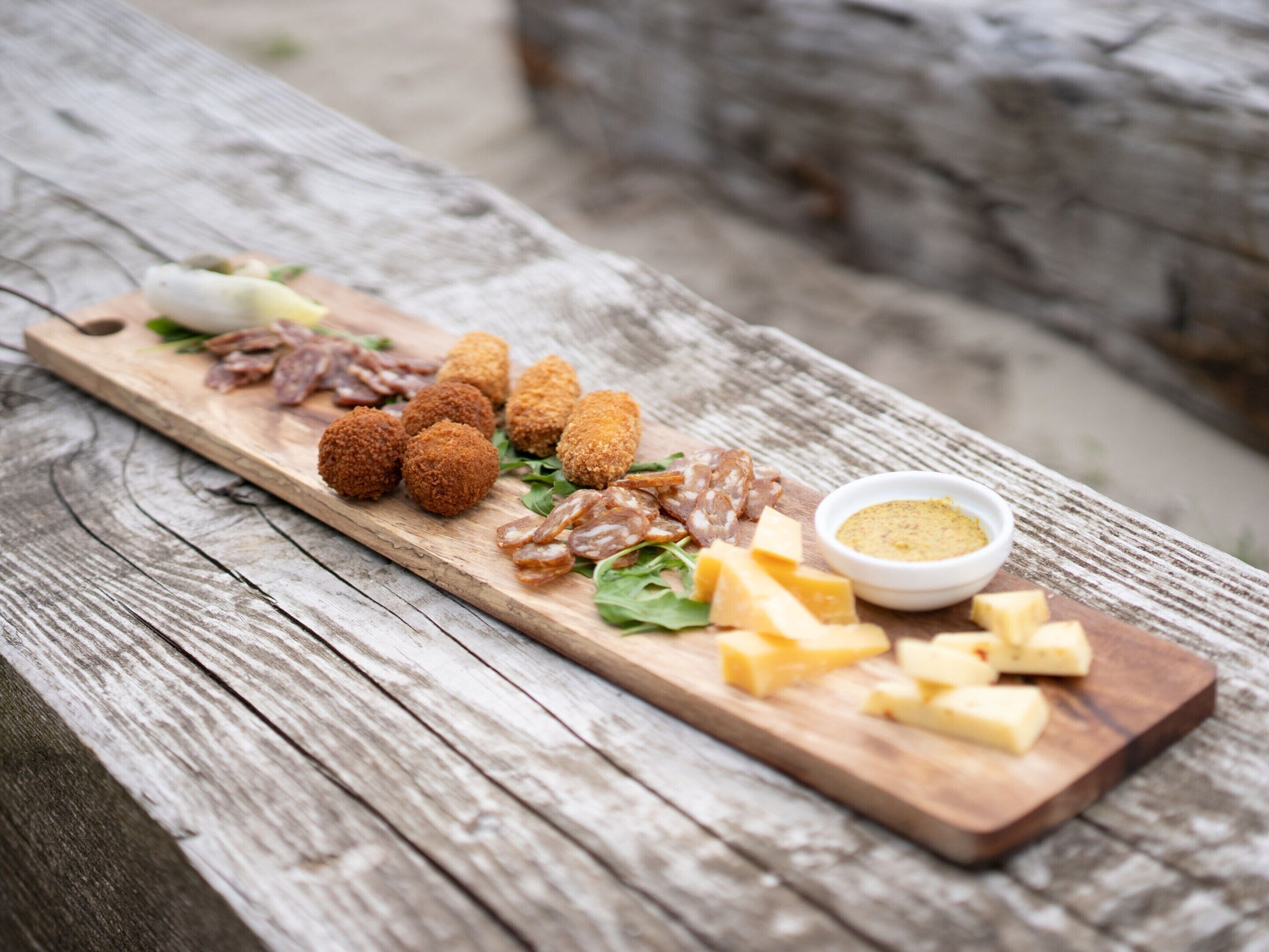 Ein Holzbrett mit verschiedenen Speisen, darunter Kroketten, Käse und Nüsse, serviert mit einer Soße.