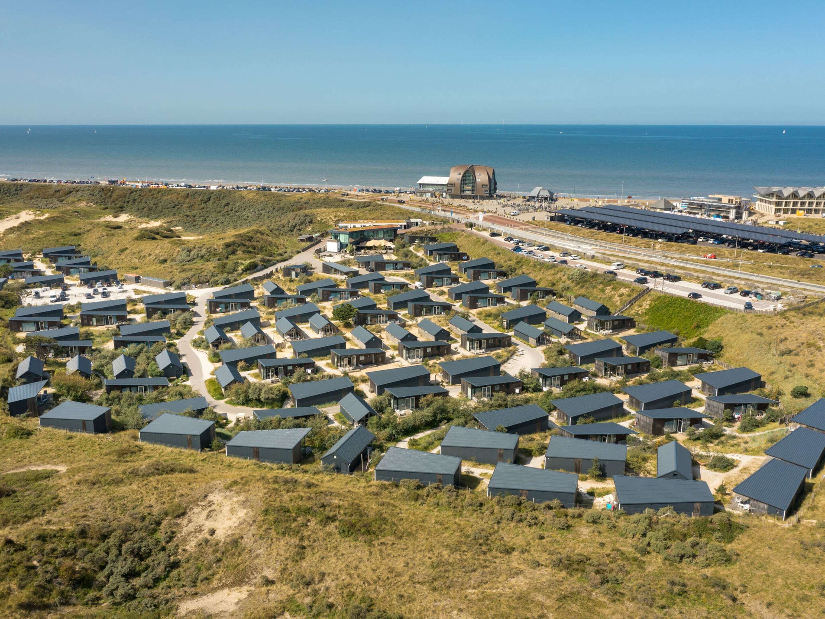 Une station balnéaire dotée de chalets modernes aux toits sombres, disposés selon un plan en réseau, d’un port de plaisance et d’un grand bâtiment à toit incurvé situé près de la plage.