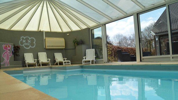 En swimmingpool med glasloft, hvide liggestole og udsigt til et hus.