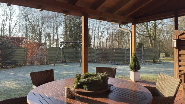 A covered patio with a round wooden table, wicker chairs, and a small potted plant. - Ferienhaus in Lindern mit Garten & Terrasse (Lindern)