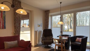 A living room with a red sofa, a glass coffee table, a brown armchair, and a black sofa. There are two pendant lights, a radiator, and a window with blinds. - Ferienhaus in Lindern mit Garten & Terrasse (Lindern)