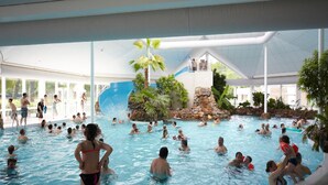 Une piscine intérieure dotée de plusieurs couloirs de nage, d’un toboggan aquatique et d’une formation rocheuse entourée de palmiers.