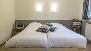 Een tweepersoonsbed met witte lakens, twee bedlampjes, een nachtkastje met boeken en een stoel met een bordje.