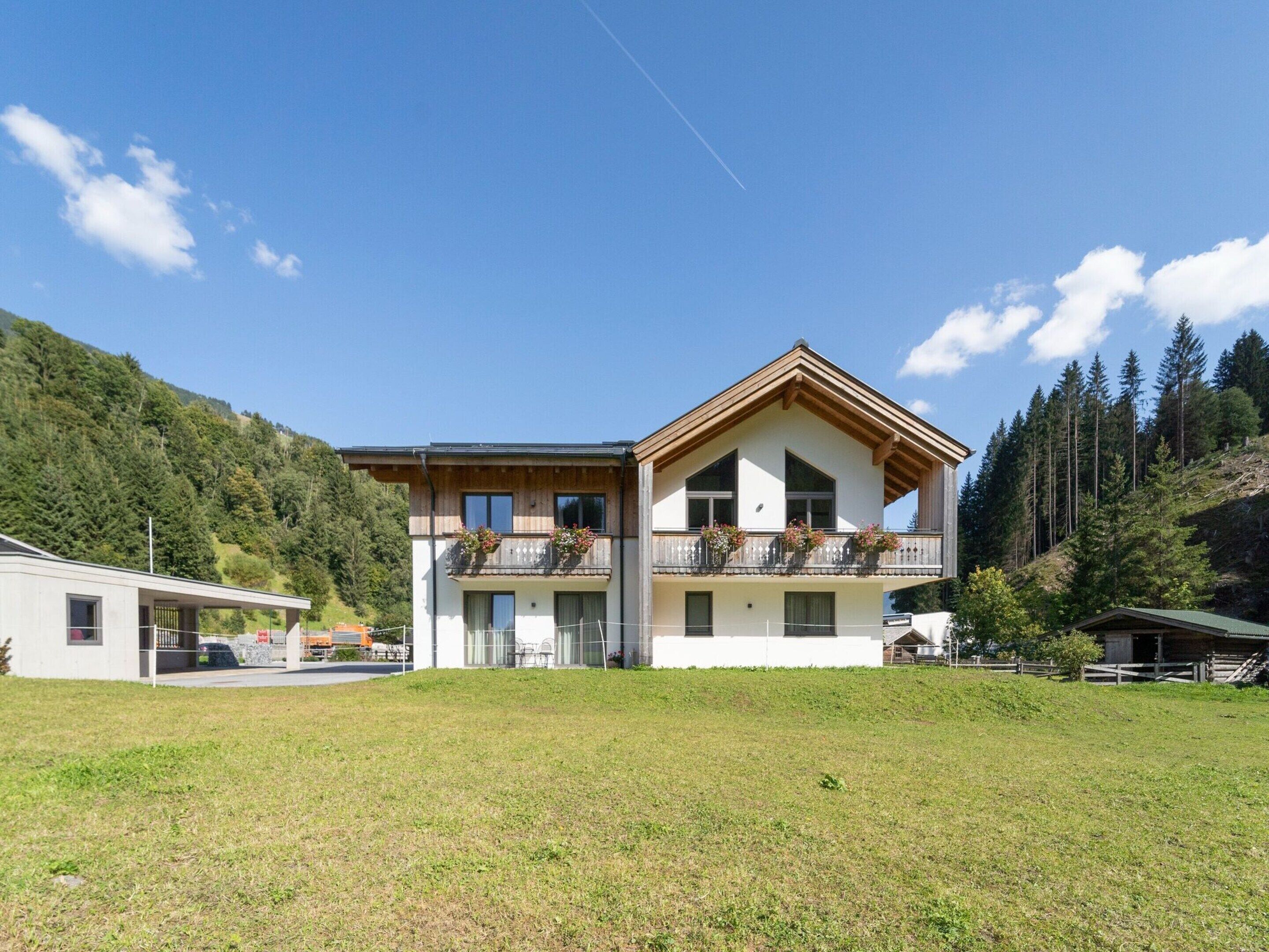 A two-story chalet with a balcony, surrounded by a grassy field and forested hills.