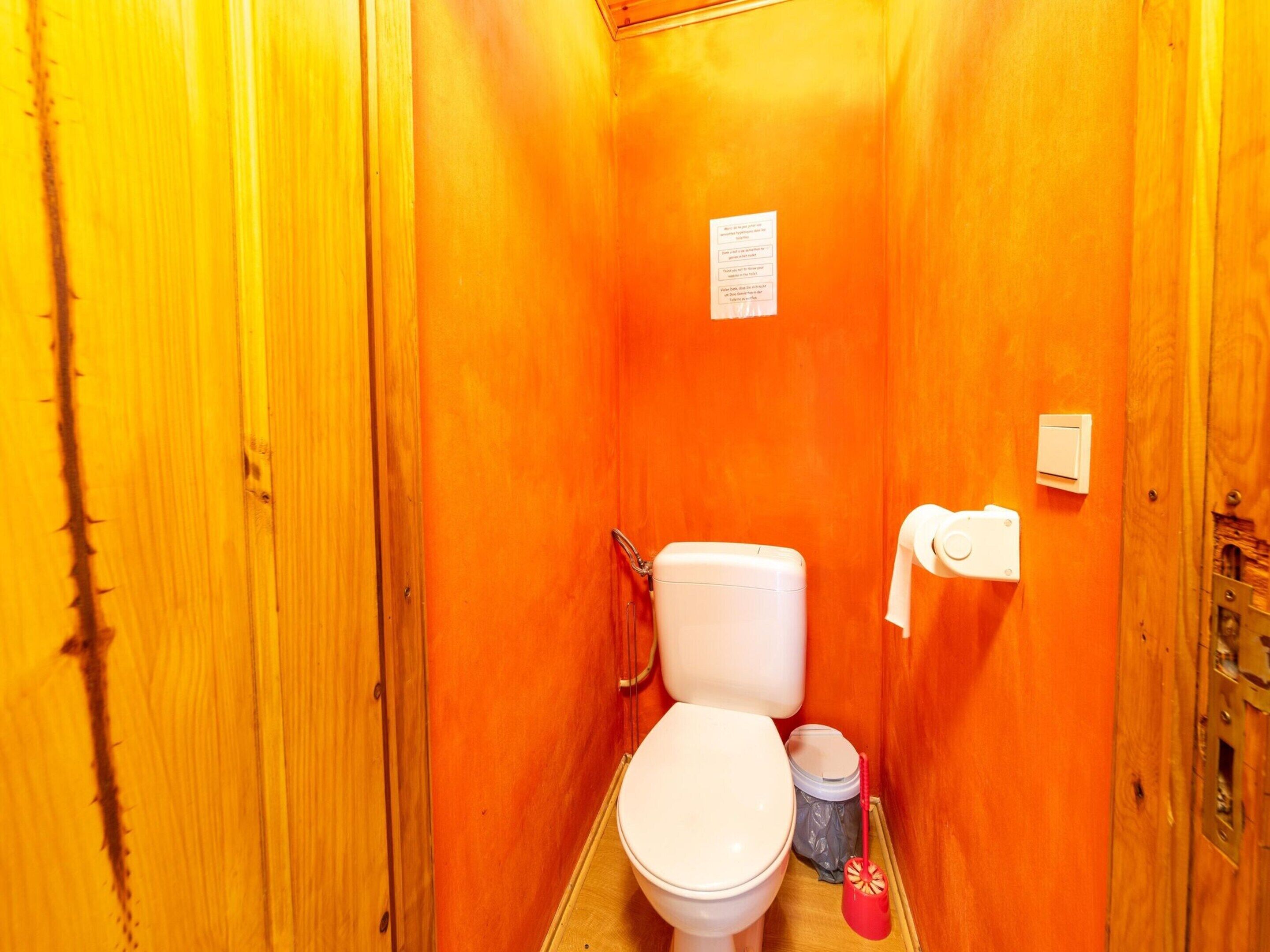 Une petite salle de bain aux murs en bois, avec des toilettes blanches, une poubelle et un déboucheur rouge.