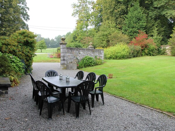 A gravel patio with a table and chairs, surrounded by lush greenery and a stone wall.