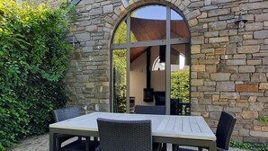 An outdoor patio with a stone wall, a large arched window, and a table with chairs. - Scenic Cottage in Rahier (Stoumont)