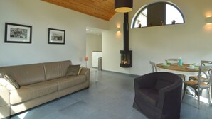 A modern living room with a high ceiling, a built-in fireplace, a dining area with a table and chairs, and two framed pictures on the wall. - Scenic Cottage in Rahier (Stoumont)