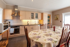 Een keuken met houten kasten, een roestvrijstalen afzuigkap, een eettafel met een bloemrijk tafelkleed en stoelen.