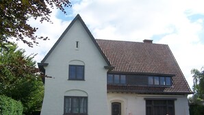Une maison à deux étages, dotée d’un toit à forte pente, d’un petit porche à l’avant et d’un jardin où poussent des fleurs rouges.