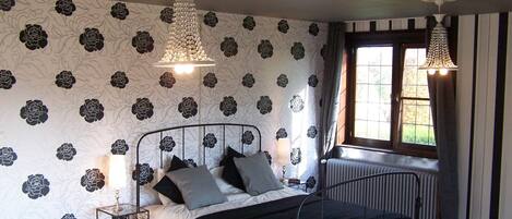 A bedroom with a bed, a radiator, a window with curtains, and a patterned wallpaper.