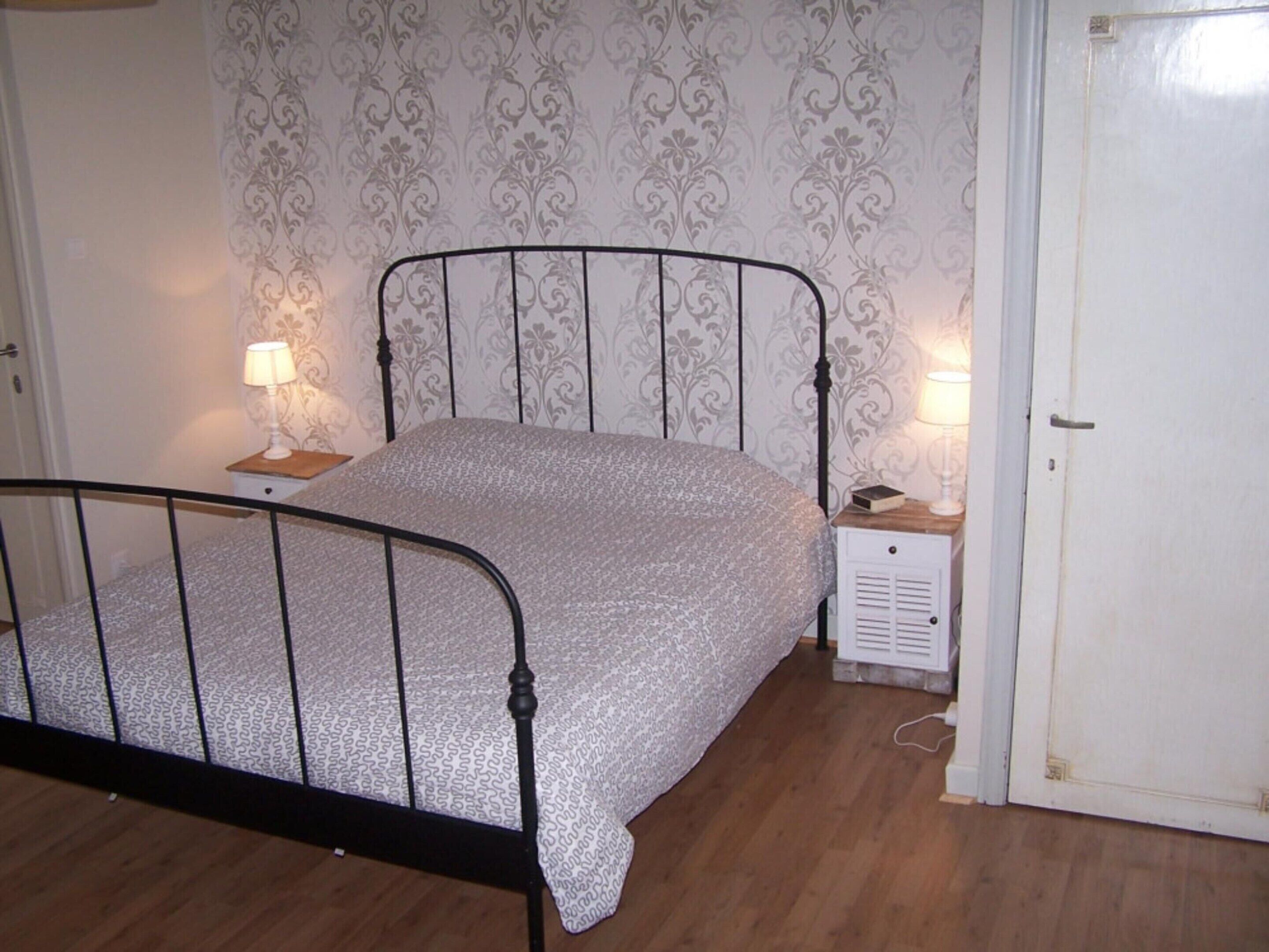 A bedroom with a metal bed frame, patterned wallpaper, a bedside table with a lamp, and a nightstand with a book.
