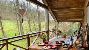 Une terrasse en bois avec une table dressée pour le repas, entourée d’une balustrade, et donnant sur une zone herbeuse avec des arbres.
