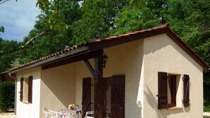 Un edificio pequeño con tejado de tejas, un porche cubierto con sillas y una mesa, y una puerta con ventana.